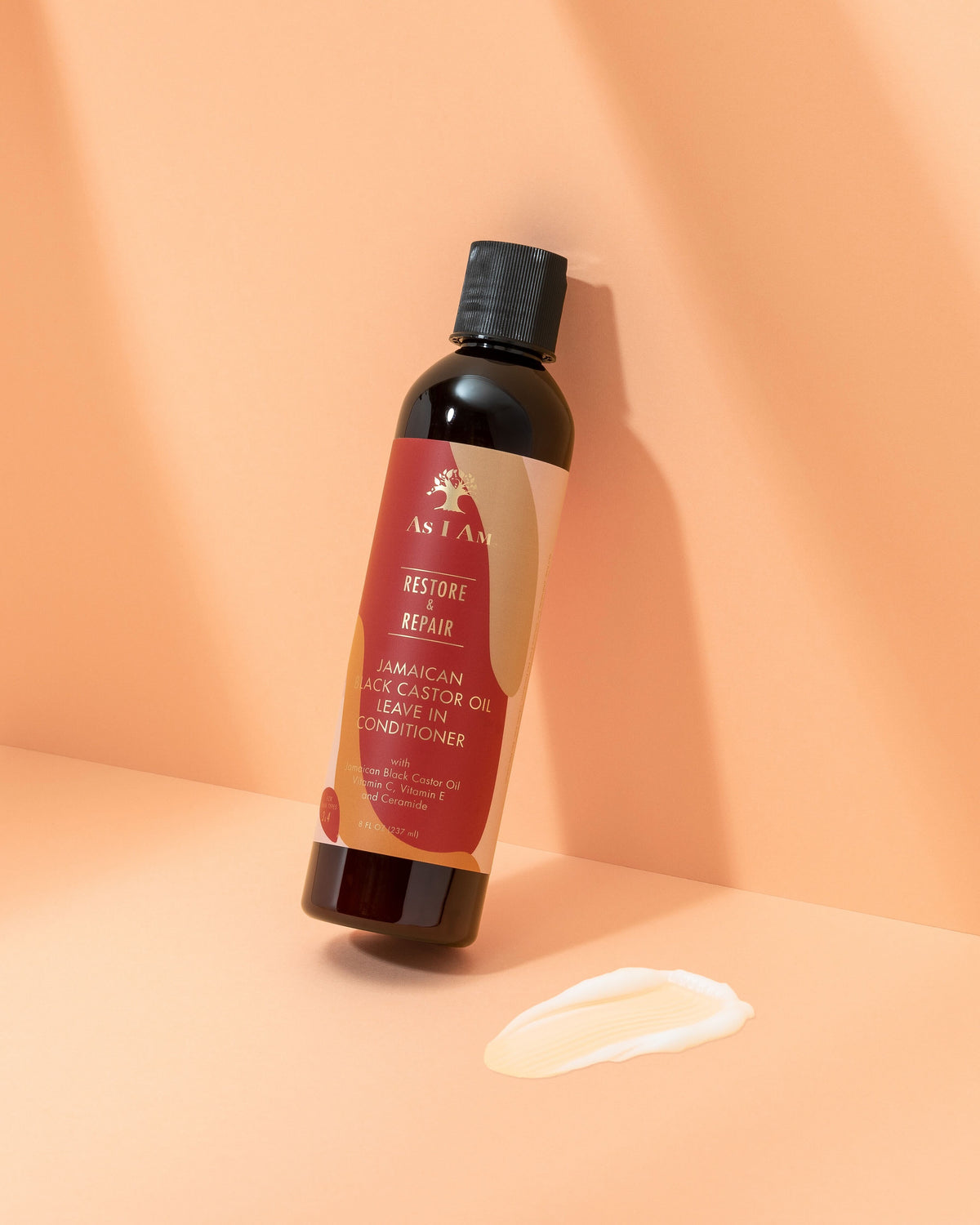 A brown plastic bottle of As I Am Jamaican Black Castor Oil Leave-in Conditioner on a light surface. The product has a creamy consistency, as shown in a small sample next to the bottle.
