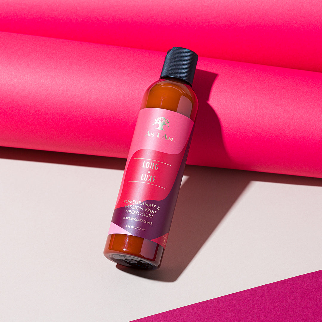 A brown bottle of As I Am Long & Luxe GroYogurt Leave-in Conditioner (237 ml) with a pink label lying diagonally in front of a pink background.