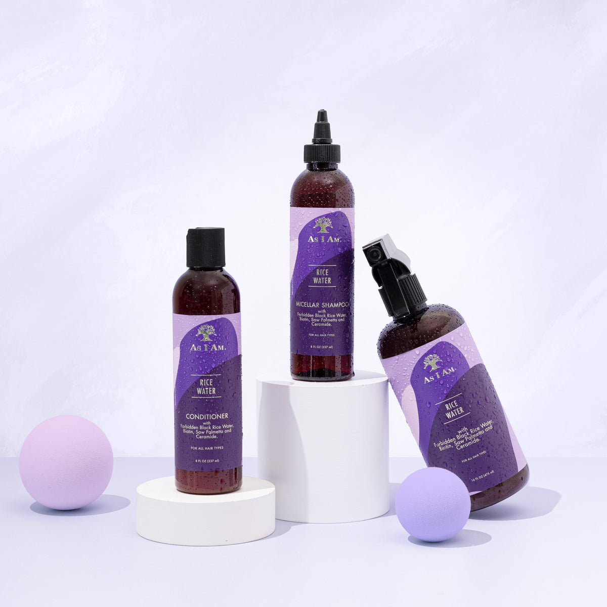 Three products from the As I Am Rice Water range: Micellar Shampoo, Conditioner and Spray, are arranged on white podiums with purple balls on a white background.
