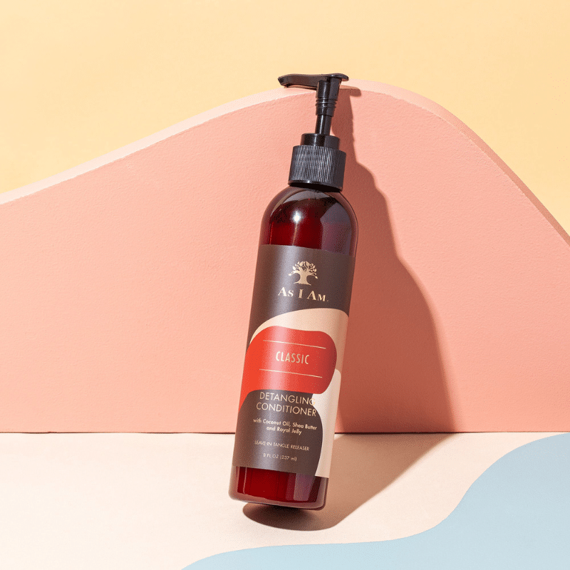 A brown bottle with a black pump stands on a pastel background. The bottle contains As I Am Classic Detangling Conditioner and has a brown label with yellow writing.