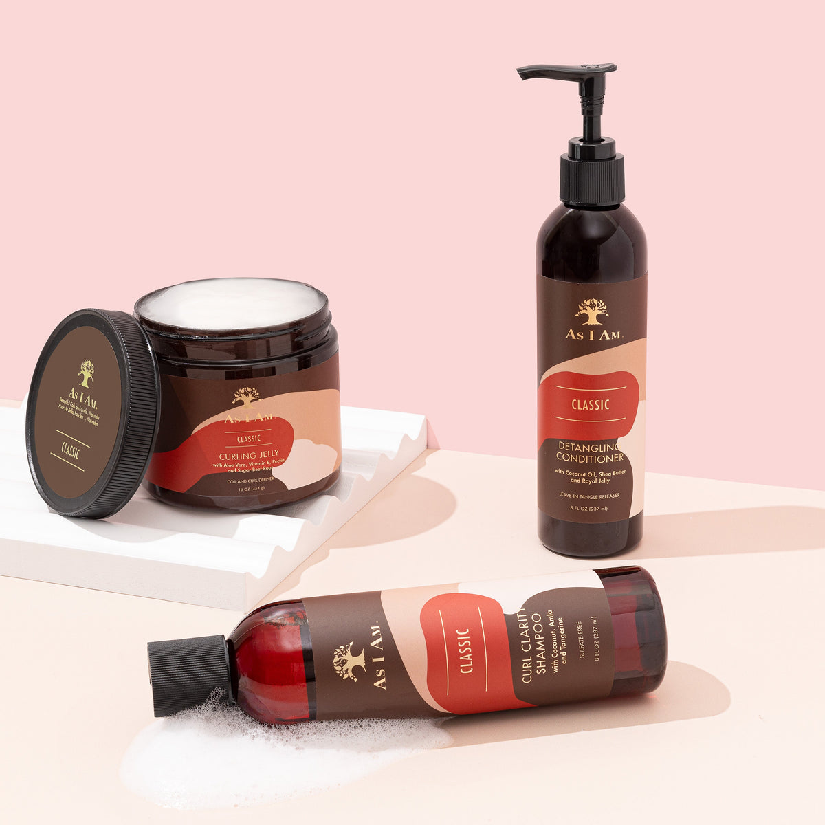 Three hair care products from As I Am stand on a light table top. From left to right you can see a small bottle of shampoo, a large jar of hair cream and a tall bottle of conditioner. The products all have a brown color and a white label with gold details.