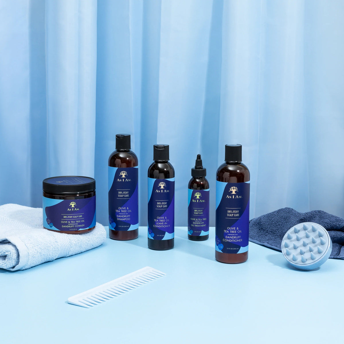 Various products from the As I Am Dry & Itchy Scalp Care range are arranged on a light blue surface with a white comb, a white washcloth and a gray scalp brush.