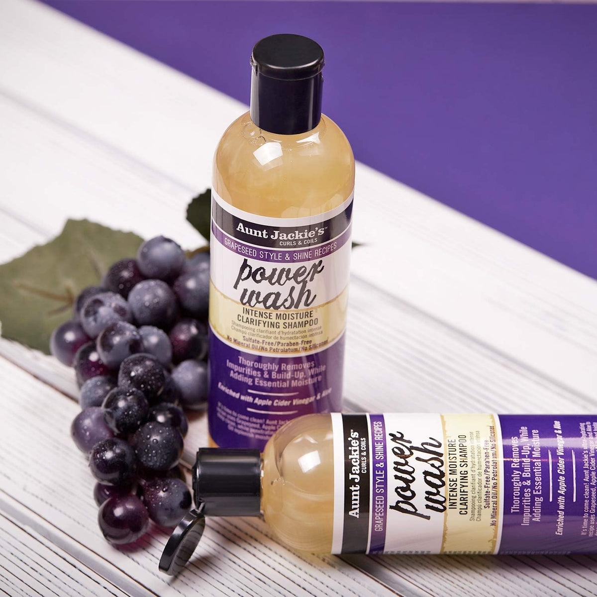 Two bottles of Aunt Jackie's Power Wash Intense Moisture Clarifying Shampoo, portrait and landscape, with a cluster of dark grapes next to it.