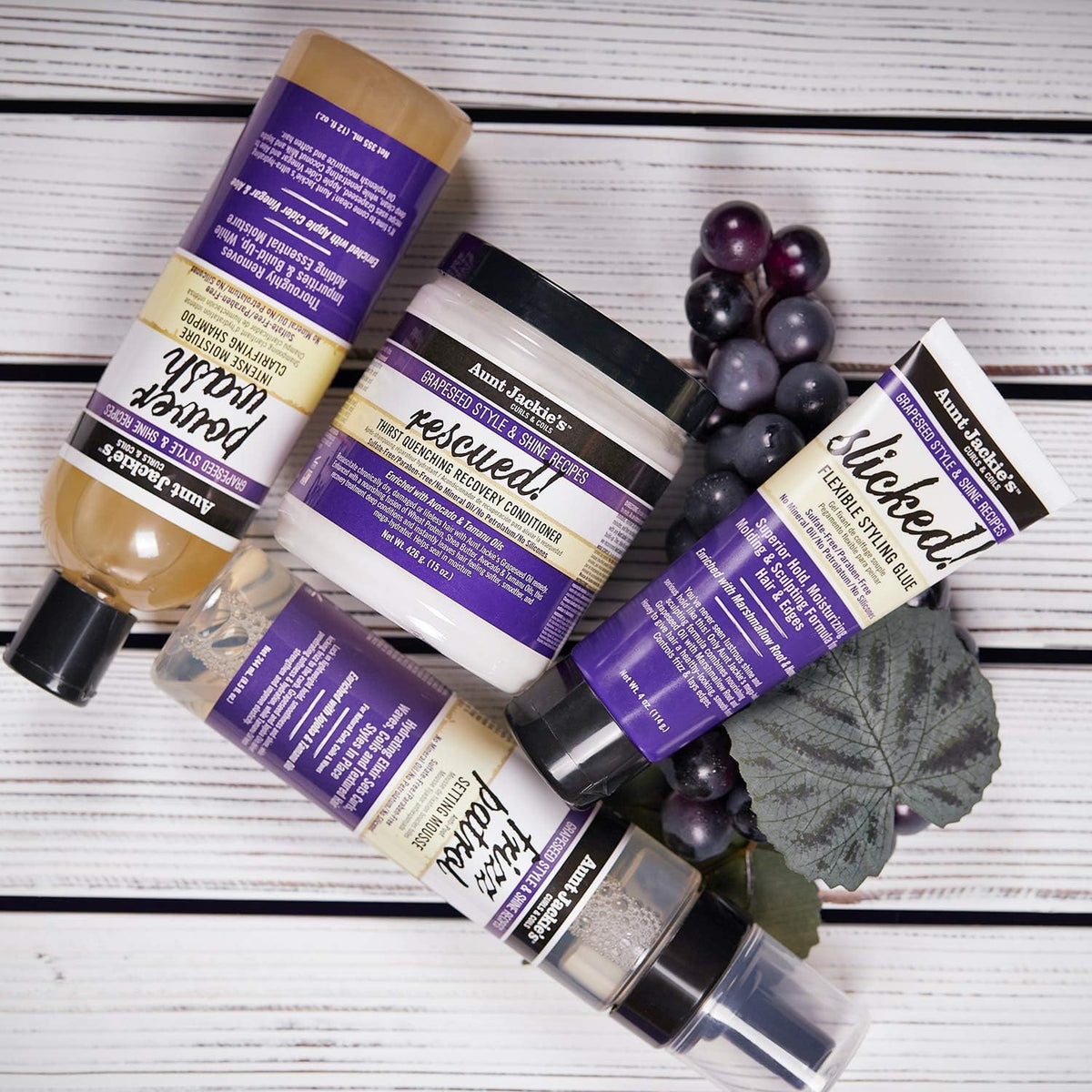 Selection of Aunt Jackie's hair products: Power Wash Shampoo, Rescued Conditioner, Slicked Styling Glue and Frizz Patrol Mousse, accompanied by dark grapes.