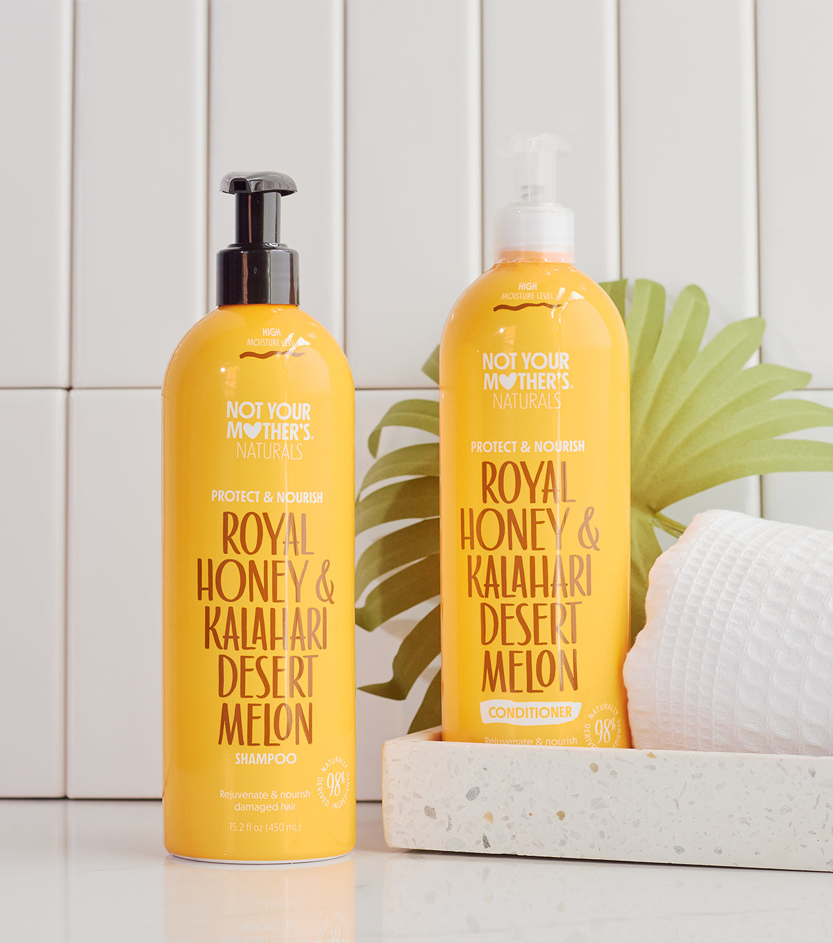 Two yellow bottles: Not Your Mother's Naturals Royal Honey & Kalahari Desert Melon shampoo (left) and conditioner (right), standing on a white surface in front of a white tile wall.