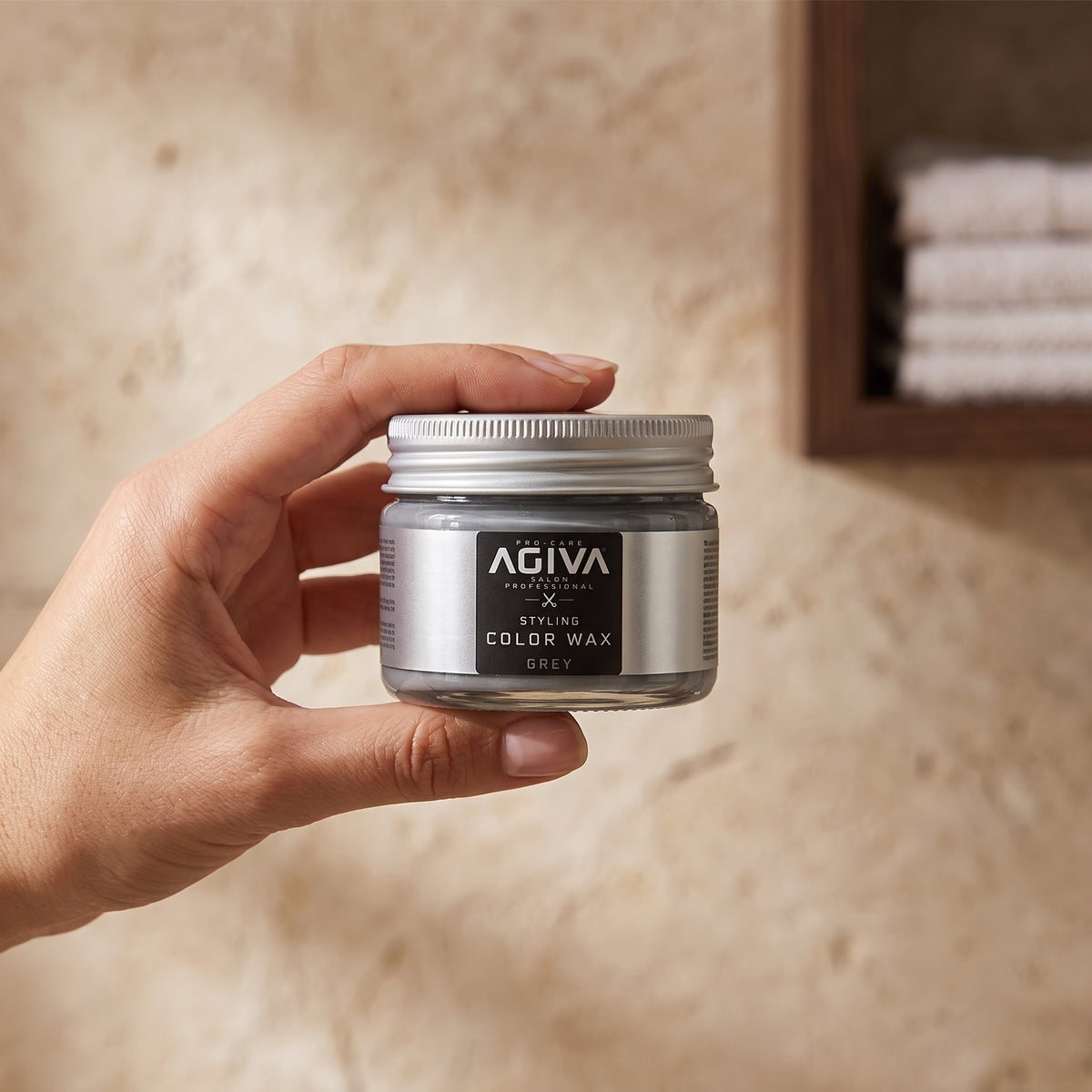 Hand holding AGIVA Pro-Care Styling Color Wax in gray colour, glass with silver lid against sand-coloured background.