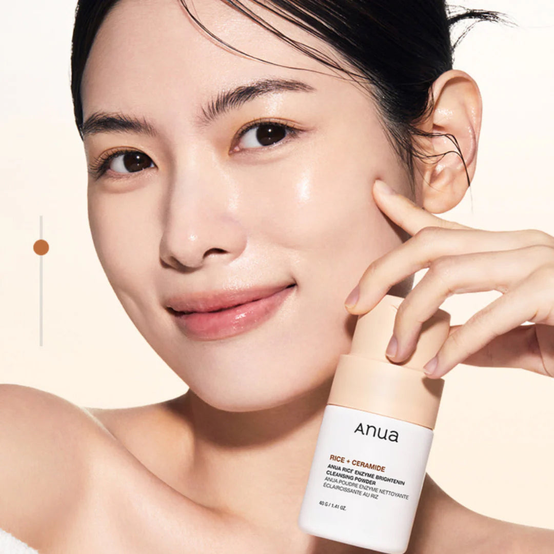 Woman with radiant skin smiles and holds Anua Rice Enzyme Brightening Cleansing Powder 40gr near her cheek.