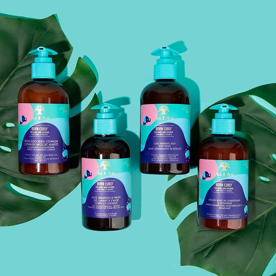 Four brown bottles of As I Am Born Curly hair care products on a blue background with green leaves. The bottles have pump dispensers and different colored labels. The products are designed for curly hair.