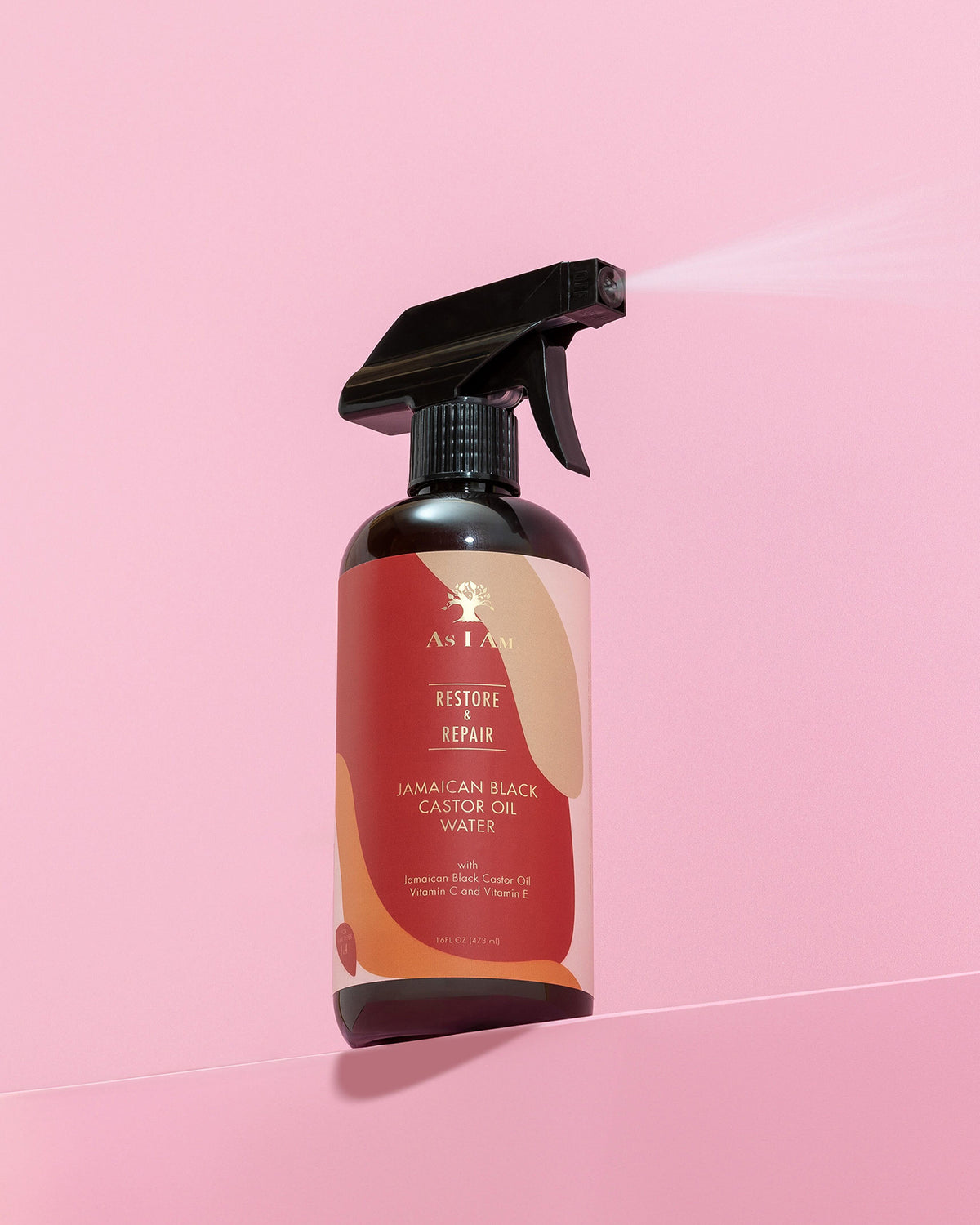 A dark spray bottle of As I Am Jamaican Black Castor Oil Water (473ml) is sprayed against a pink background showing a fine mist of the product.