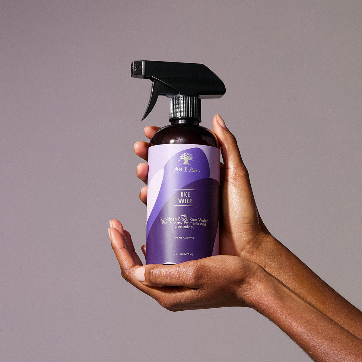 Hands holding a bottle of As I Am Rice Water Spray (475 ml) in front of a purple background.