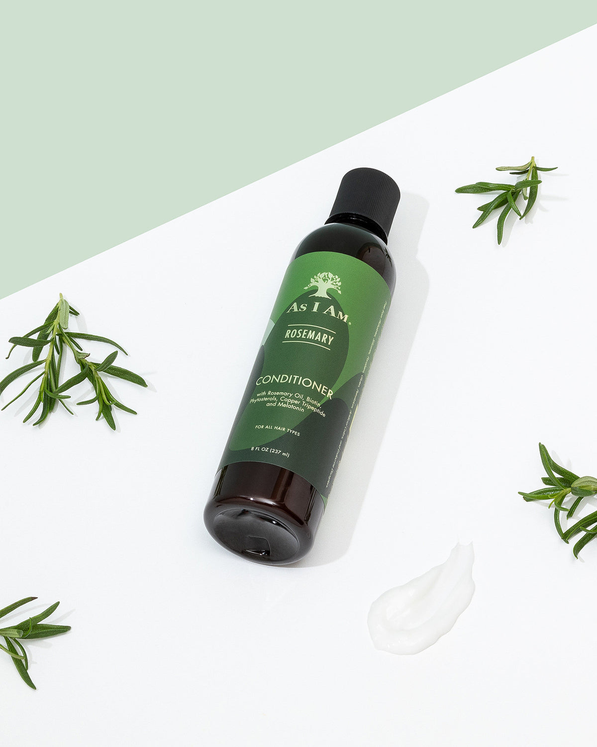 A bottle of As I Am Rosemary Conditioner (237 ml) lies diagonally on a white surface with rosemary sprigs scattered around and a dollop of conditioner.