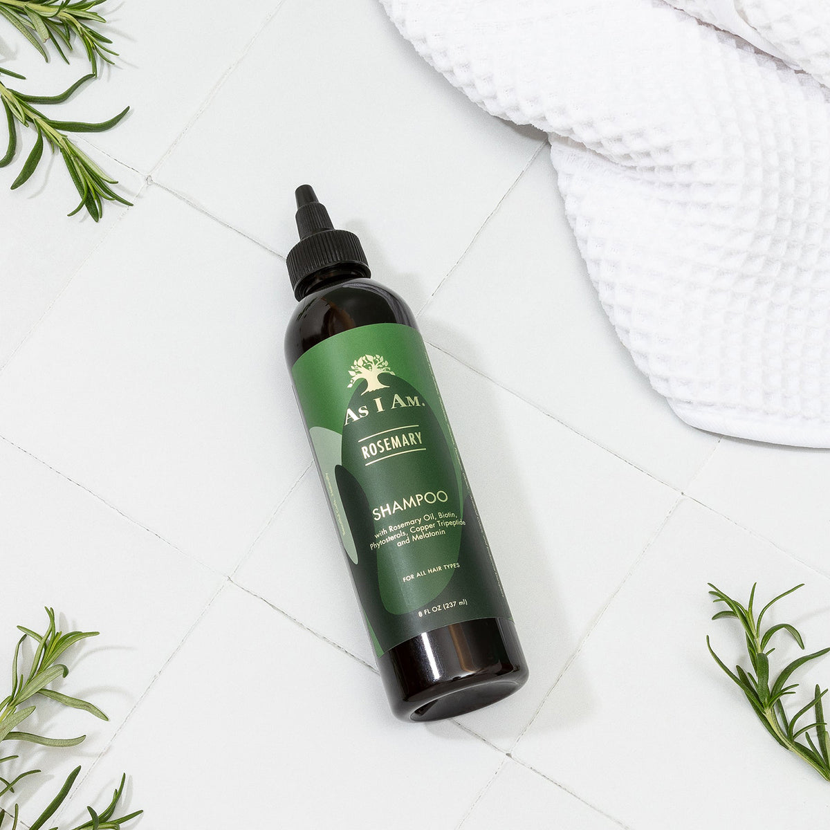 A bottle of As I Am Rosemary Shampoo (237ml) lies diagonally on a white tiled surface, surrounded by fresh rosemary sprigs and a white towel.
