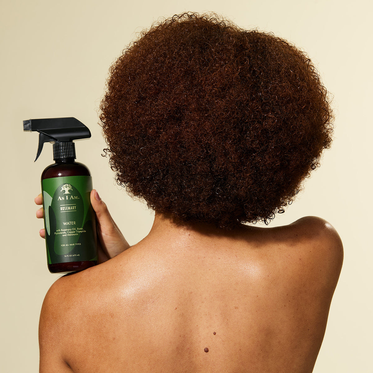 A person with curly hair holds a bottle of As I Am Rosemary Water Spray (475 ml) and is about to spray the product into their hair.