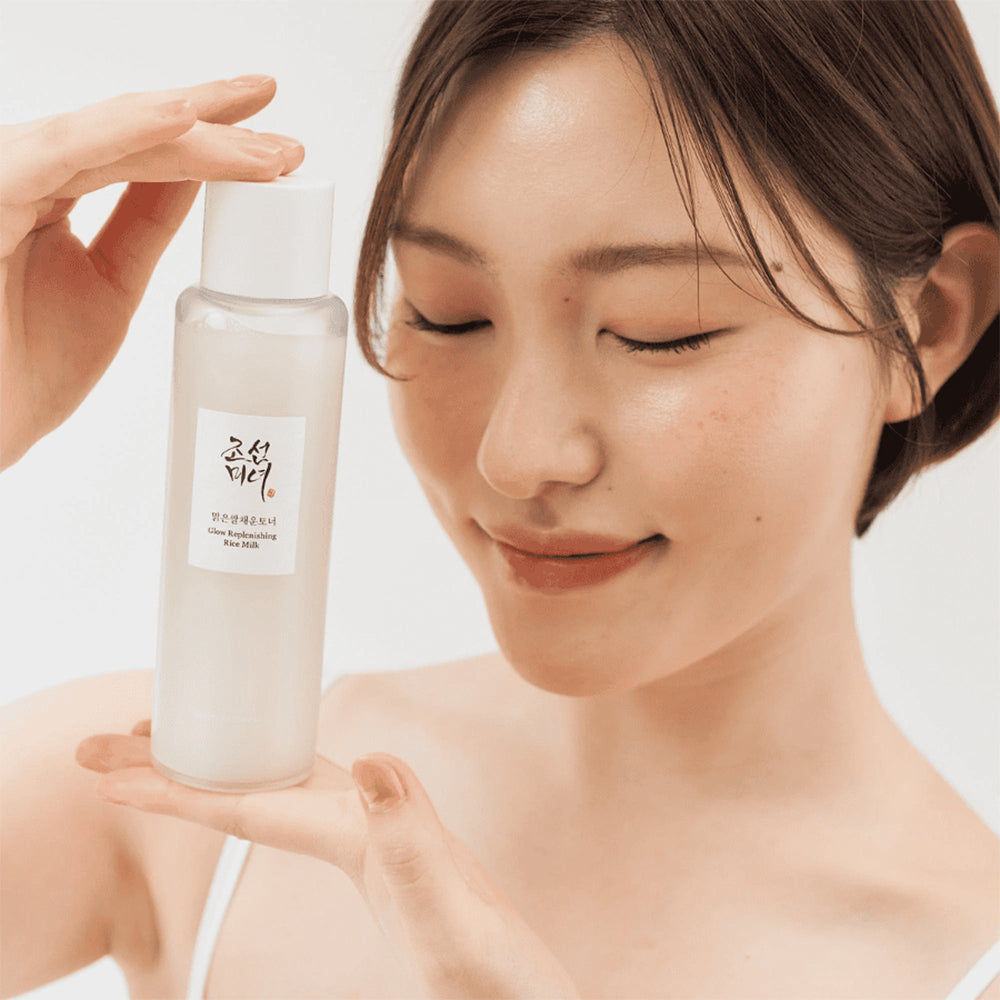 Woman with glowing skin holds Beauty Of Joseon Glow Replenishing Rice Milk 150ml in front of her face and smiles with closed eyes.