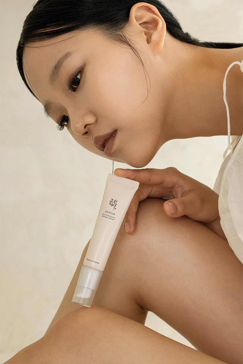Woman with glowing skin holds Beauty of Joseon Revive Eye Serum Ginseng + Retinal 30ml in a neutral setting.