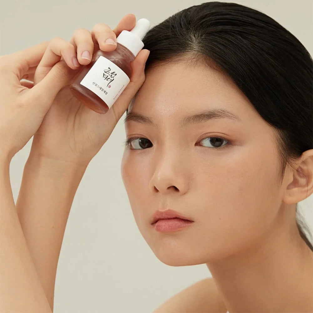 Woman with glowing skin holds Beauty of Joseon Revive Serum: Ginseng + Snail Mucin 30ml up to her forehead.