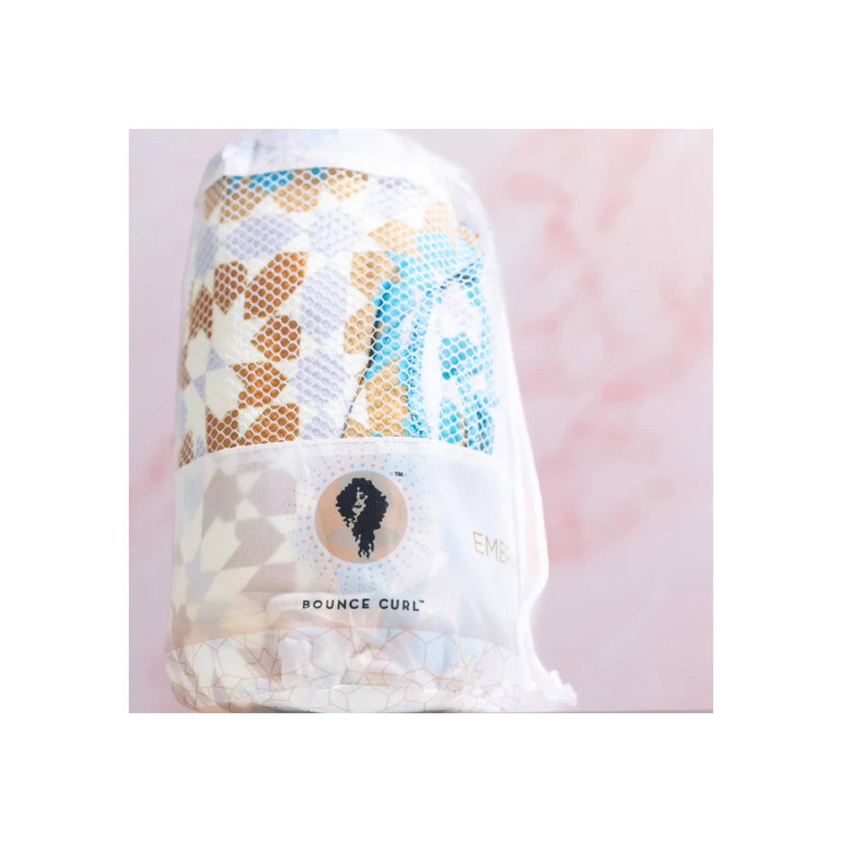 Colorful Bounce Curl Micro-fiber Towel in mesh bag with geometric pattern and logo in front of pink background.