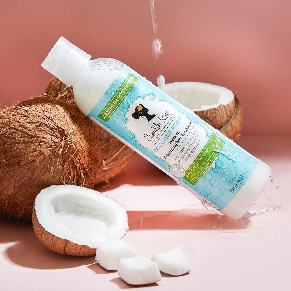 White bottle of Camille Rose Coconut Water Leave-In Treatment sits next to an open coconut with water droplets.
