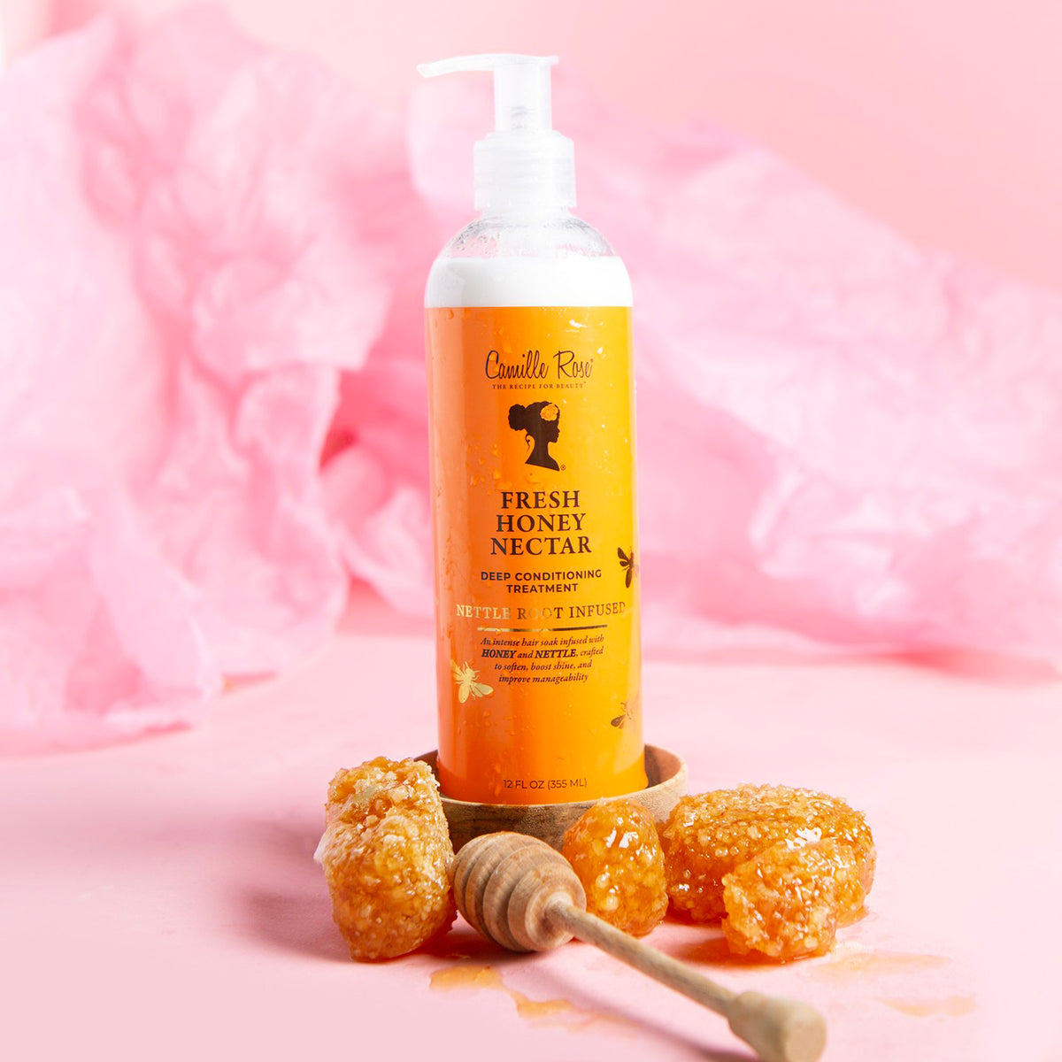 White bottle with pump of Camille Rose Fresh Honey Nectar Deep Conditioning Treatment on a pink background. Next to the bottle are honey combs and a honey dipper.
