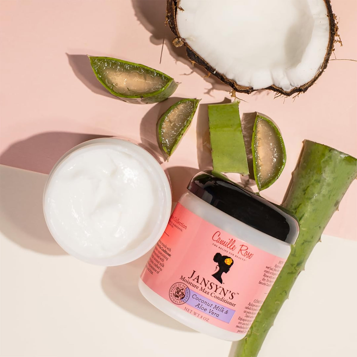 Camille Rose Jansyn's Moisture Max Conditioner in a white container with a black top. The product is shown together with natural ingredients such as coconut and aloe vera.