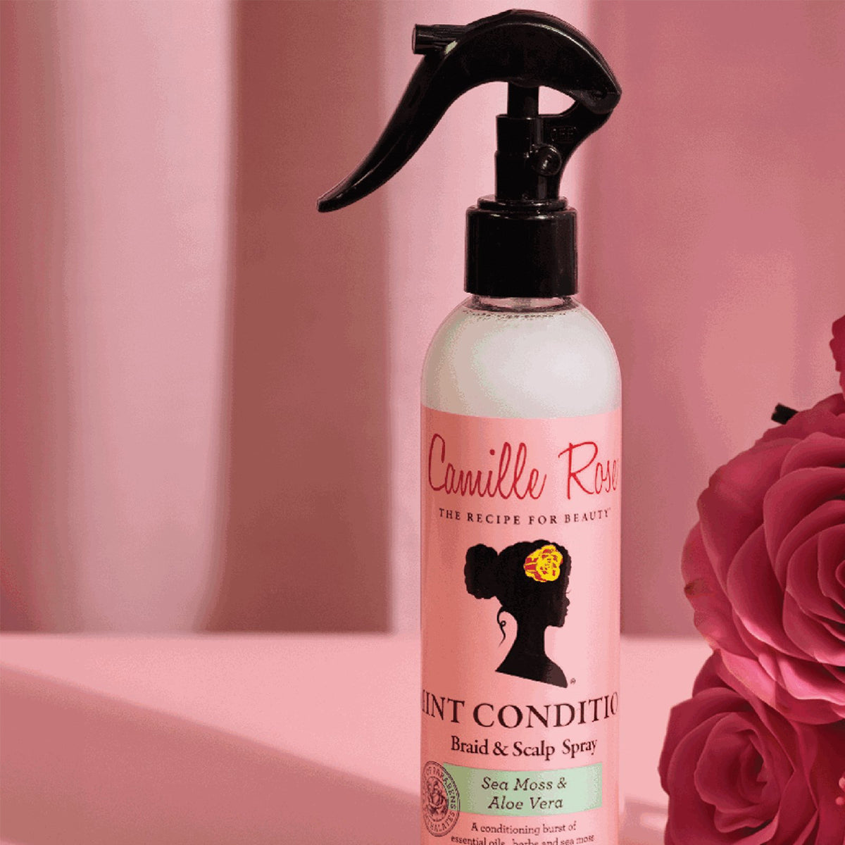 Pink spray bottle with Camille Rose Mint Condition Braid & Scalp Spray. The product is depicted against a dark background together with artificial pink roses.