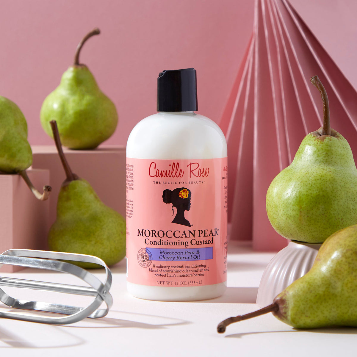 Camille Rose Moroccan Pear Conditioning Custard: White bottle with pink label, surrounded by fresh pears and a whisk, on a pink background.
