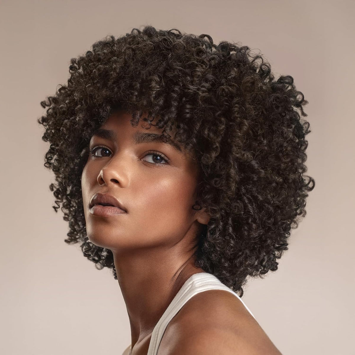 Woman with natural curly afro hairstyle and curly bangs, wearing white top against beige background.