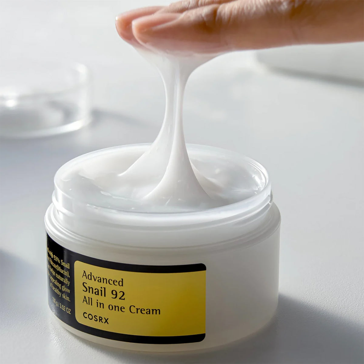Fingers lift elastic texture from open jar of Cosrx Advanced Snail 92 Cream 100gr on white table.