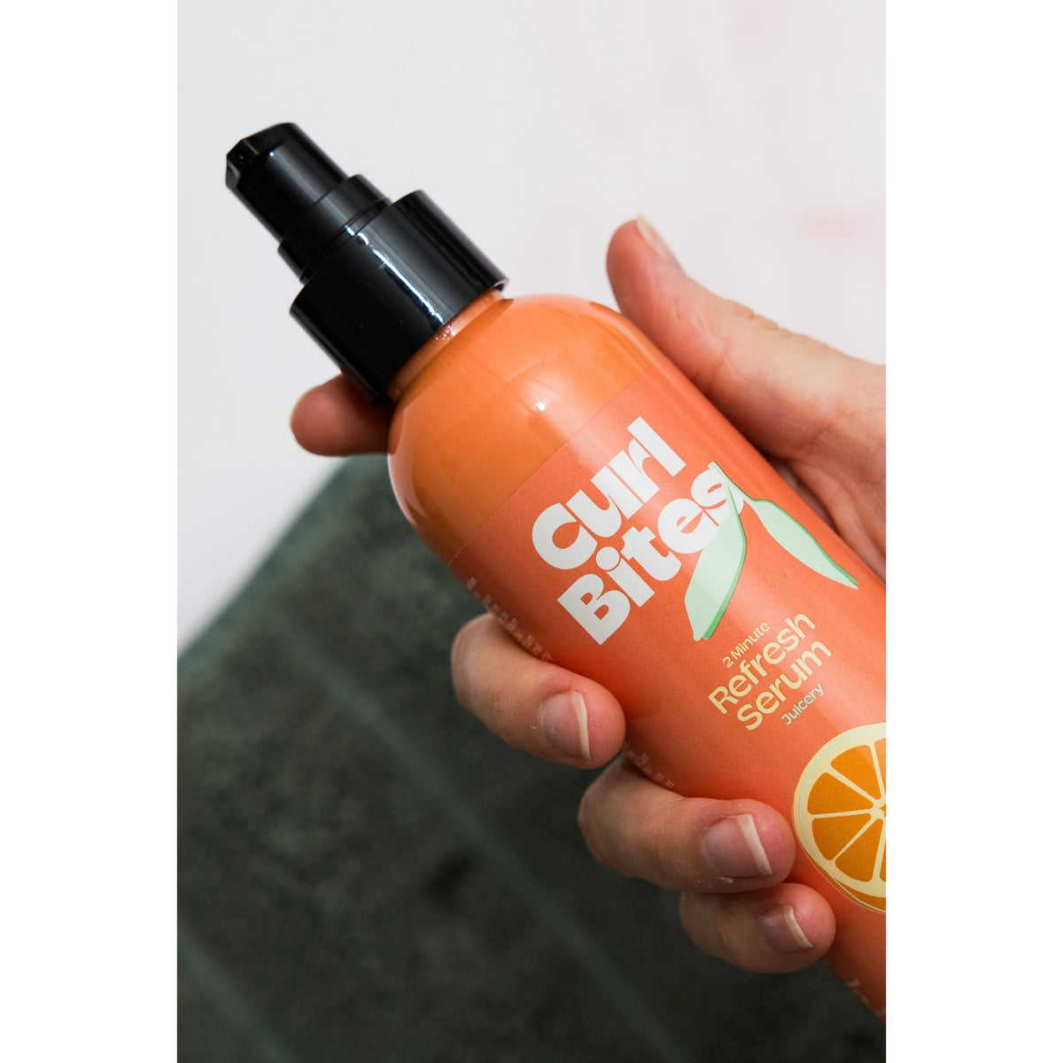 Hand holding orange Curl Bites 2 Minute Refresh Serum bottle with black pump against green background.