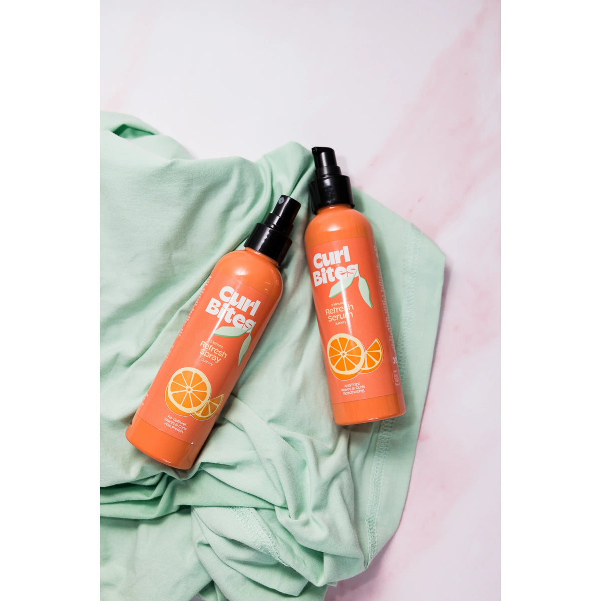 Curl Bites Refresh Spray and Refresh Serum in orange bottles with black pump caps, placed on a mint green fabric background.
