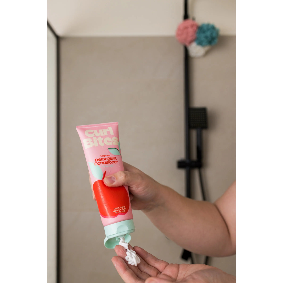 Hand squeezes Curl Bites Detangling Conditioner into pink tube with apple motif in bathroom.