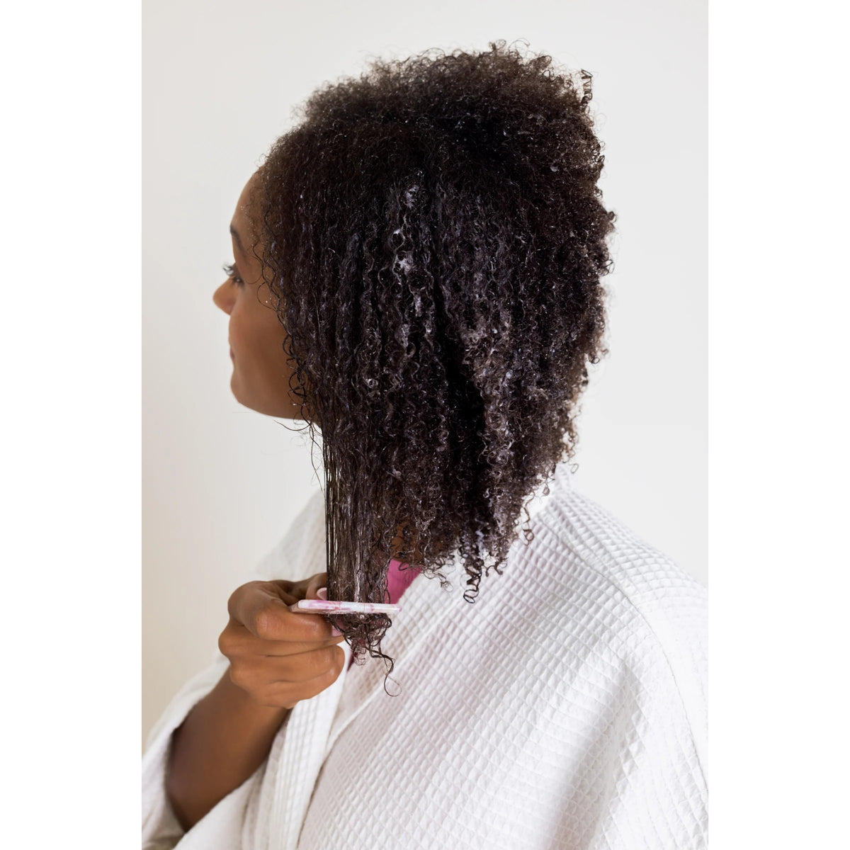 Woman with curly afro hair combs through wet curls with dental floss comb, wearing white bathrobe.