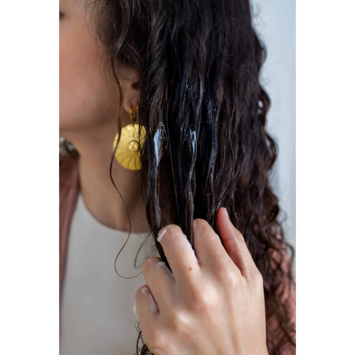 Woman with curly dark hair applies white hair mask with fingers on damp hair lengths.