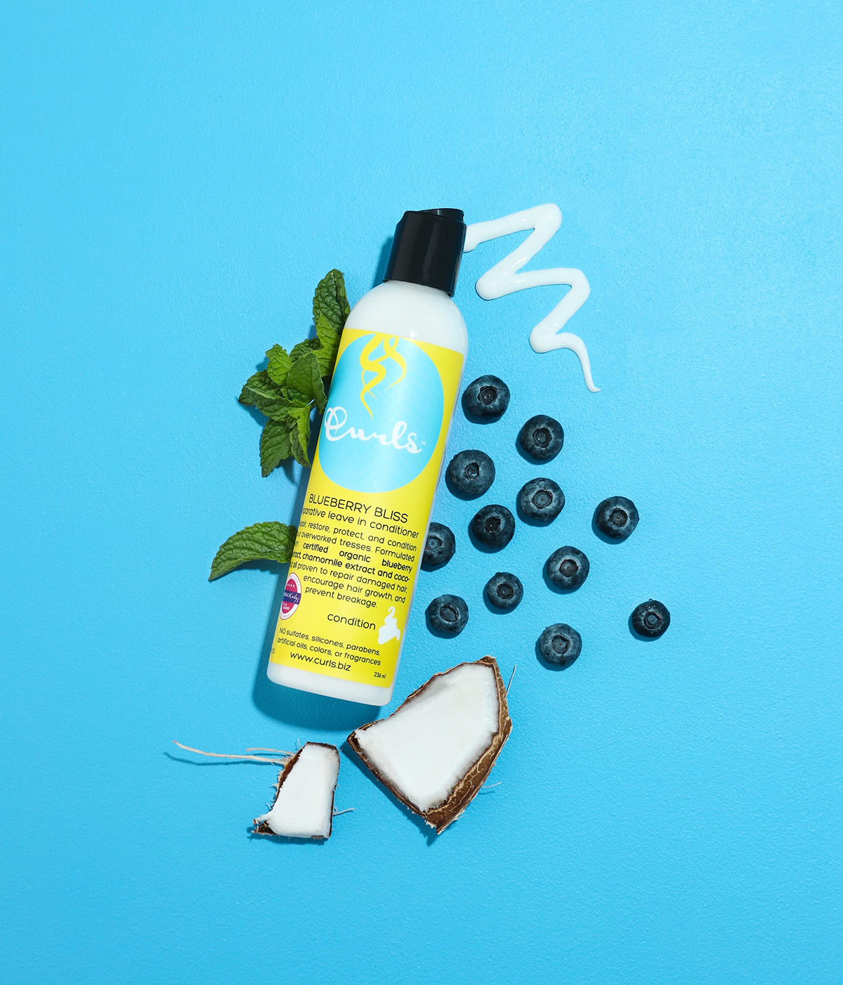 Curls Blueberry Bliss Reparative Leave-In Conditioner (236ml) presented with blueberry, mint and coconut on a pale blue background. White bottle with light blue label and black cap. Leave-in conditioner to repair damaged hair.