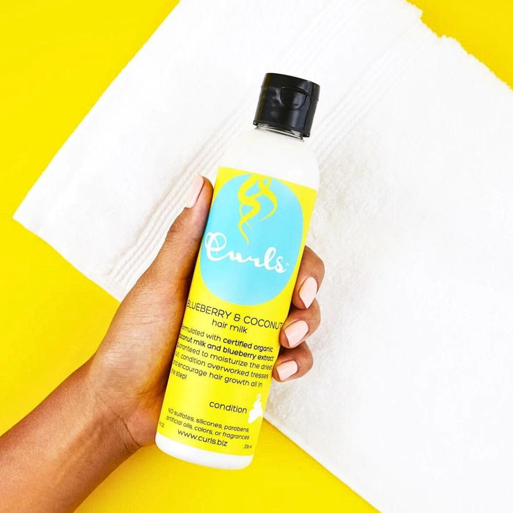 A hand holds a bottle of Curls Blueberry & Coconut Hair Milk against a white background.