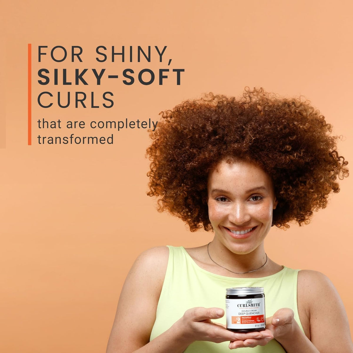Woman with auburn curls smiles and holds Curlsmith Double Cream Deep Quencher for shiny, silky curls.