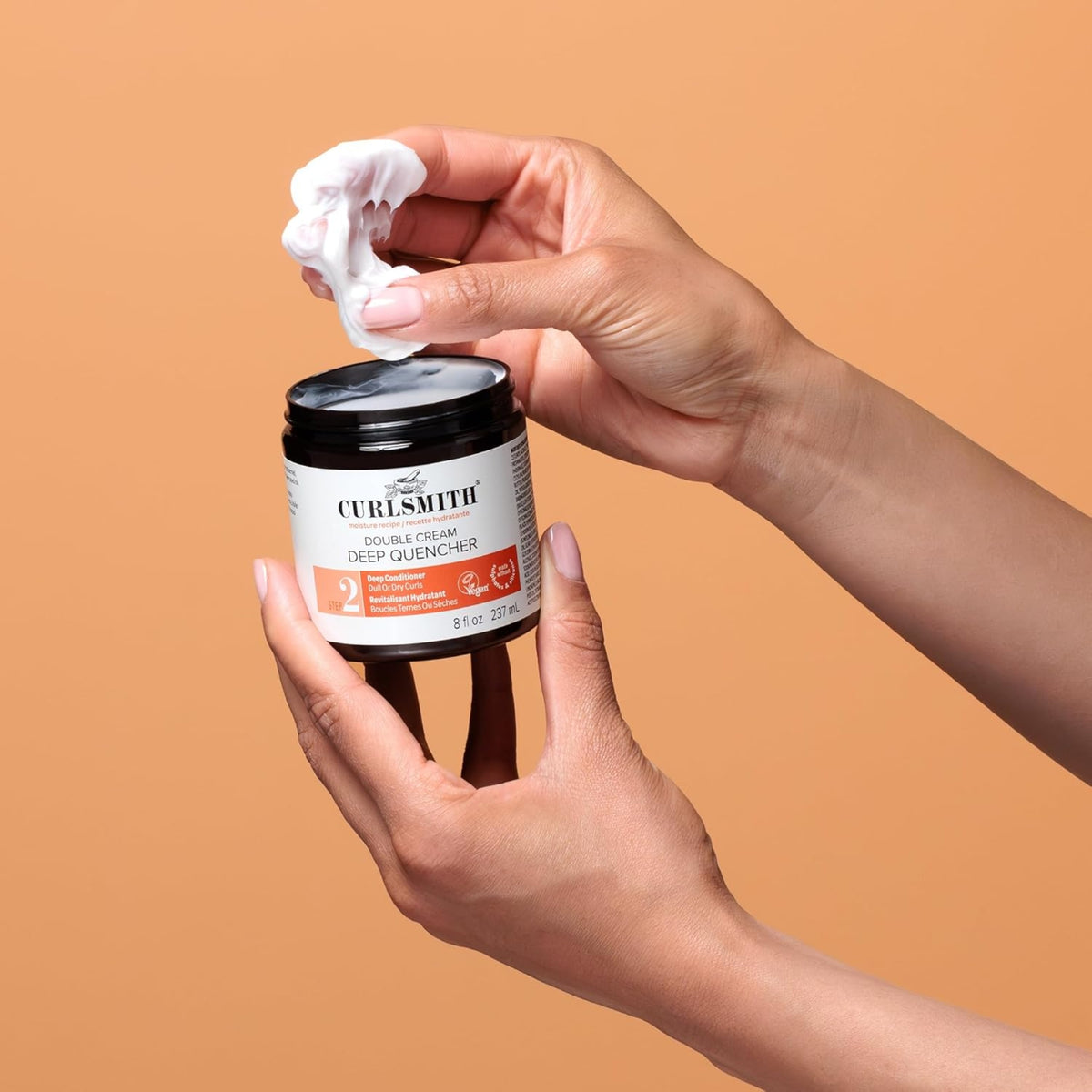 Hand picks up creamy Curlsmith Double Cream Deep Quencher hair treatment from black jar against orange background.