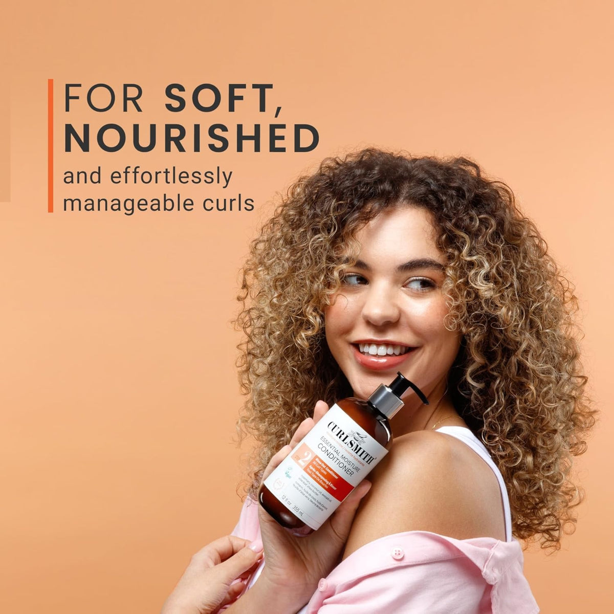 Woman with soft, nourished curls holds Curlsmith Essential Moisture Conditioner up to her shoulder and smiles.