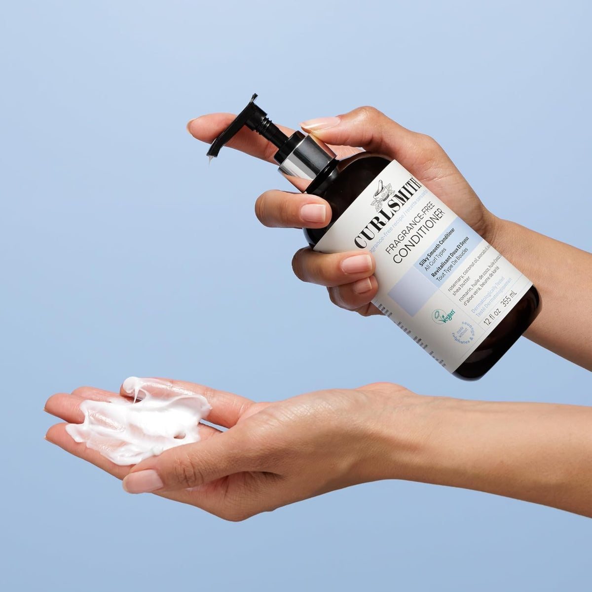 Hand holds Curlsmith Fragrance-Free Conditioner and pumps white cream into the other palm against blue background.