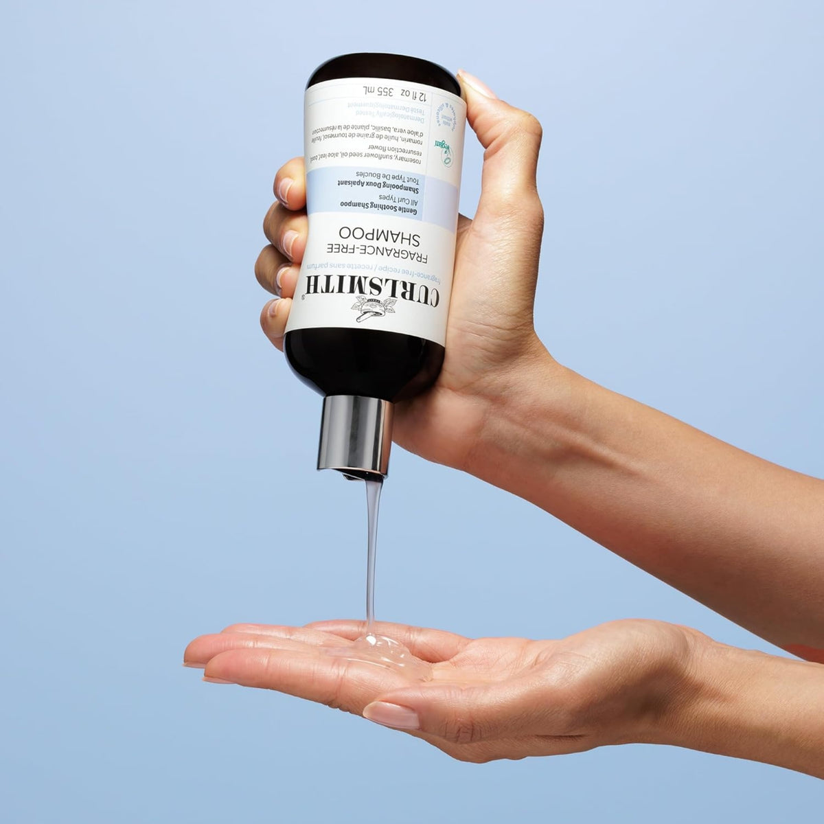 Curlsmith Fragrance-Free Shampoo is poured from the bottle into the open palm against a light blue background.