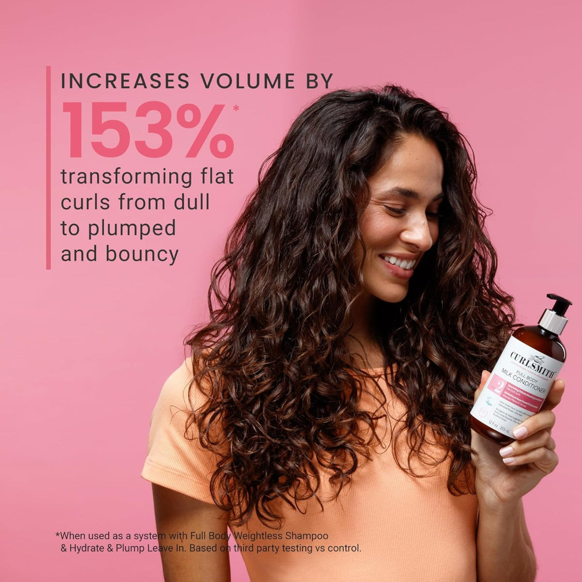 Woman with voluminous dark curls smiling and holding Curlsmith Full Body Milk Conditioner in front of pink background.