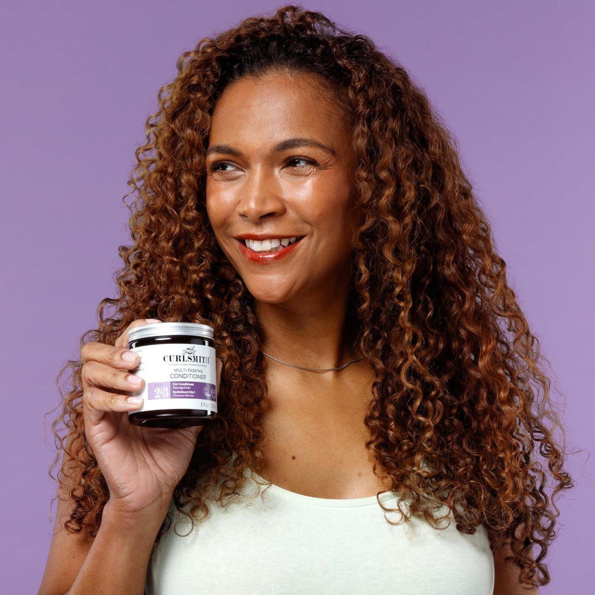 Woman with curly hair smiling and holding a can of Curlsmith Multi-Tasking Conditioner in front of purple background.