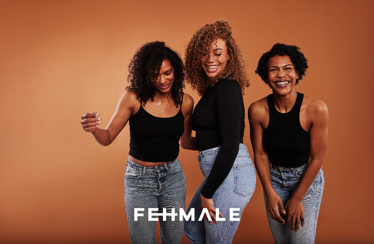 Three women with naturally curly hair in black tops and jeans smile and laugh in front of orange background, Fehmale.