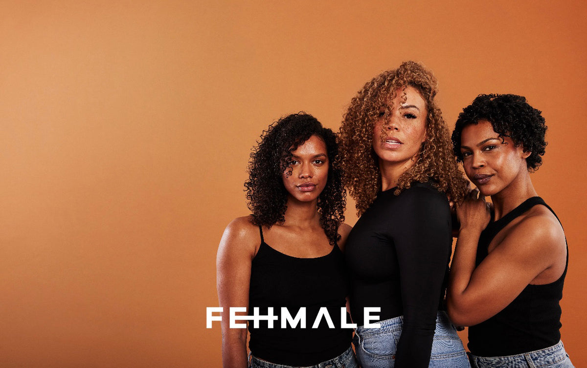 Three women with natural curls pose in front of orange background wearing black clothes, promoting Fehmale.