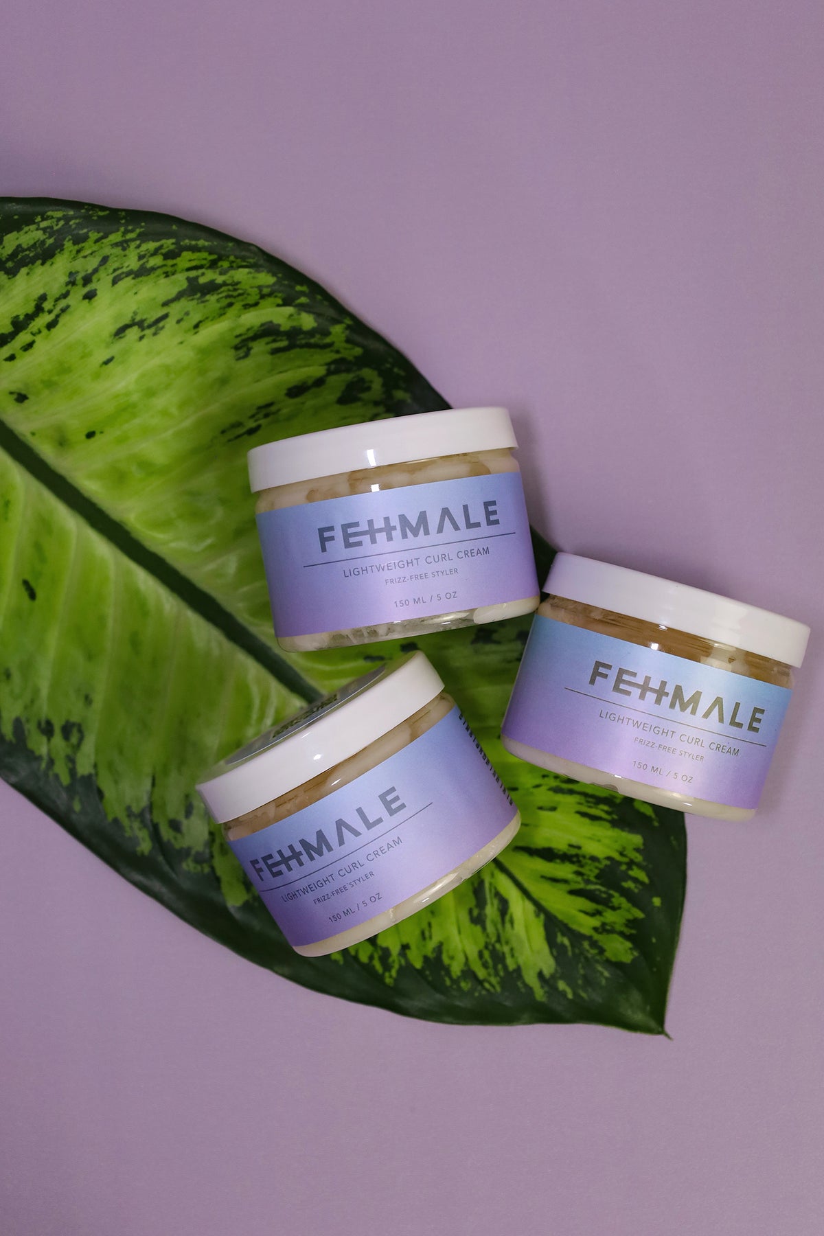 Three Fehmale Lightweight Curl Cream 150ml jars on green leaf decoration against purple background, perfect for curly hair.