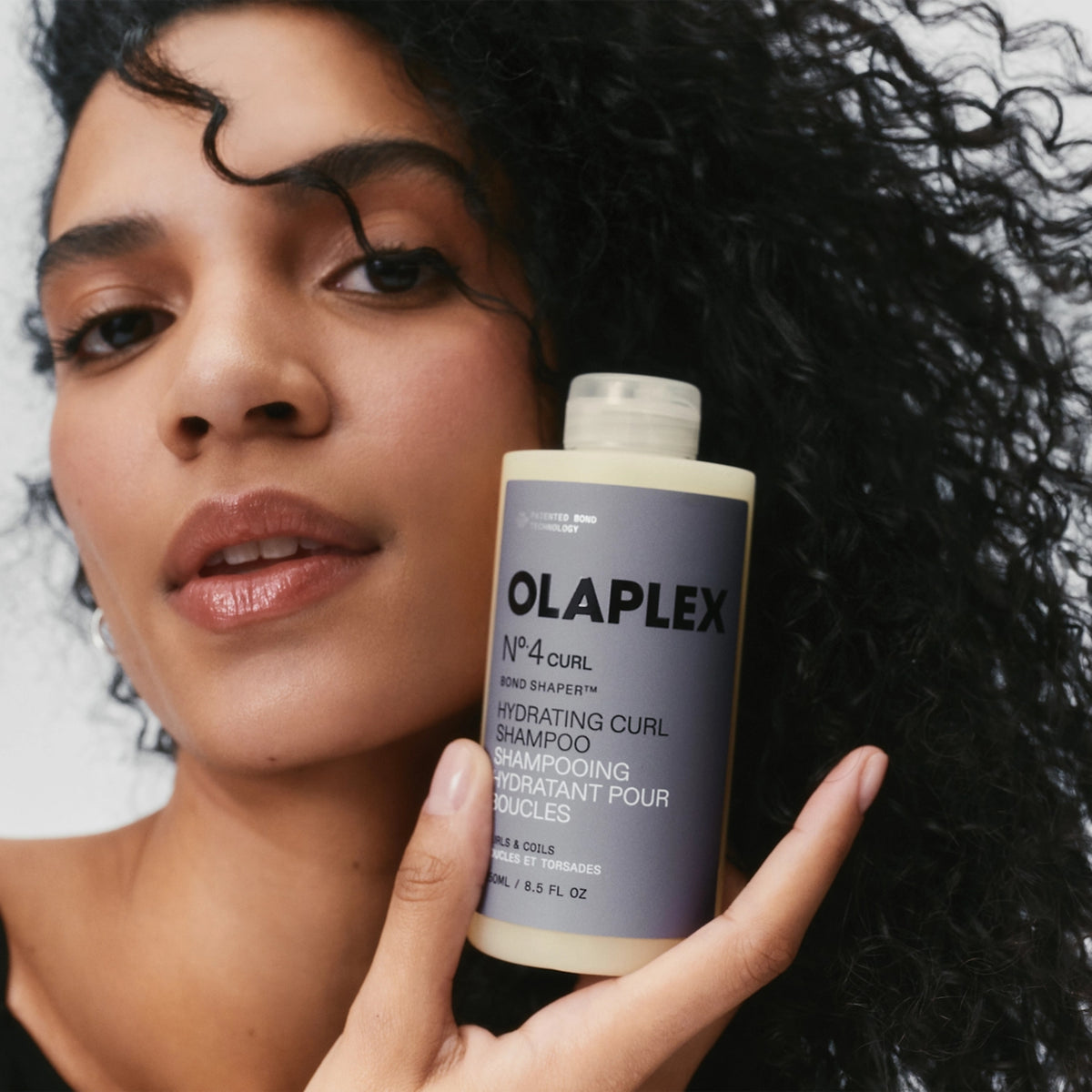 Woman with dark curls holds Olaplex Nº.4 Curl Bond Shaper hydrating curl shampoo close to face.