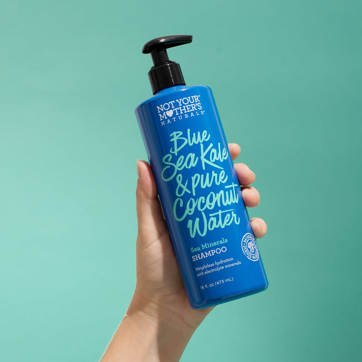 Close-up of a hand holding a blue bottle of Not Your Mother's Naturals Blue Sea Kale & Pure Coconut Water Sea Minerals Shampoo against a turquoise background.