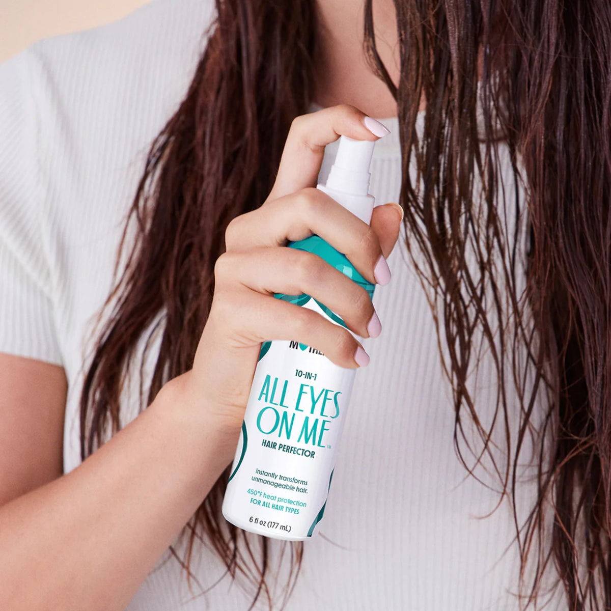 Woman with long wet brown hair sprays All Eyes On Me Hair Perfector heat protection into her hair.