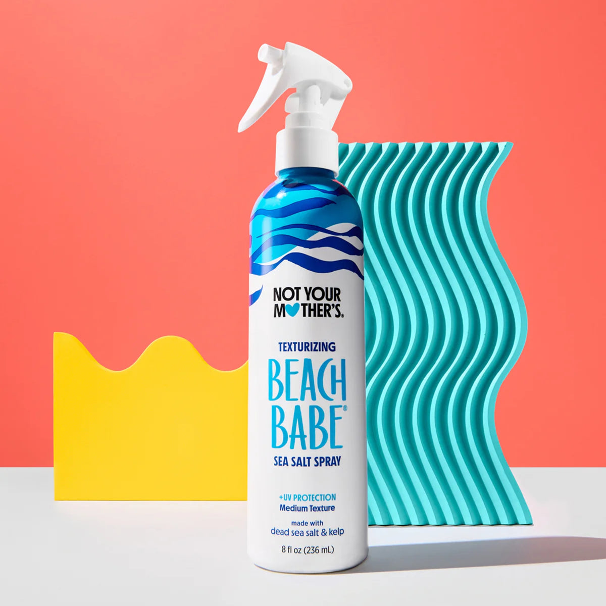 Not Your Mother's Beach Babe sea salt spray in front of turquoise wave pattern on coral red background.