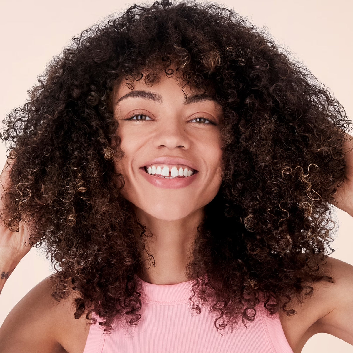 Smiling woman with full natural curly hair and pink tank top against beige background.